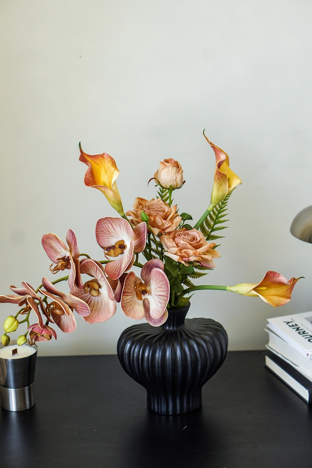 Faux floral arrangement with peach orchids, garden roses, and orange calla lilies in a matte black ribbed vase on a dark desk