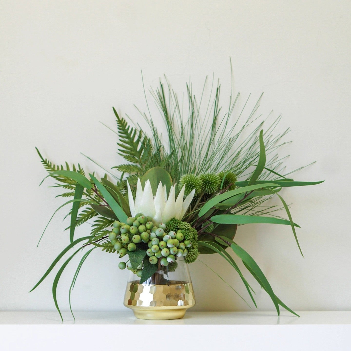 Tropical green floral arrangement with white King Protea and green berries in a matte gold honeycomb glass vase on a white shelf