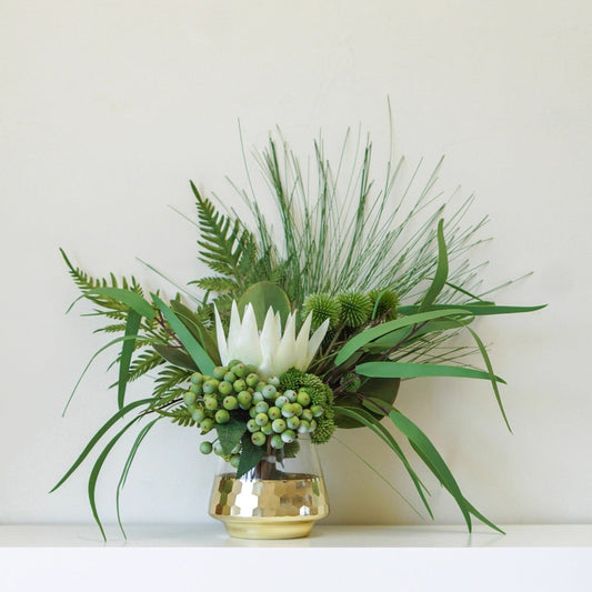 Tropical green floral arrangement with white King Protea and green berries in a matte gold honeycomb glass vase on a white shelf
